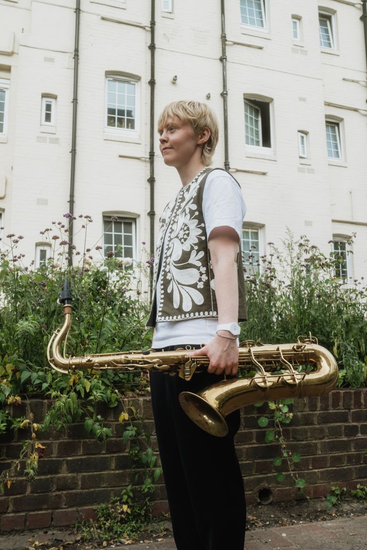 Person stood by a wall in front of a multi story house, holding saxophone